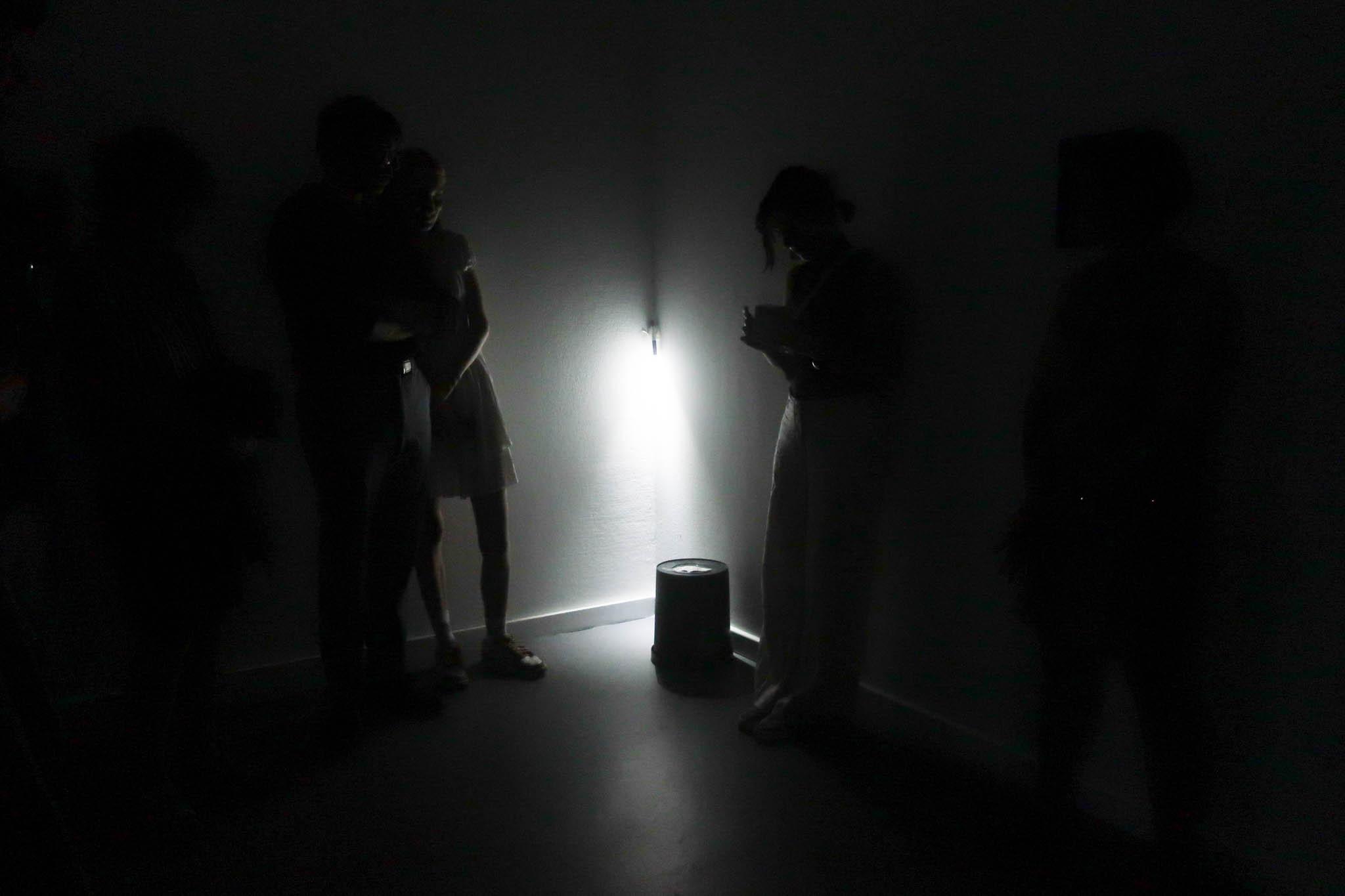 A dark, desaturated space, with only a single white light in a corner illuminating dim figures surrounding an upturned bucket.